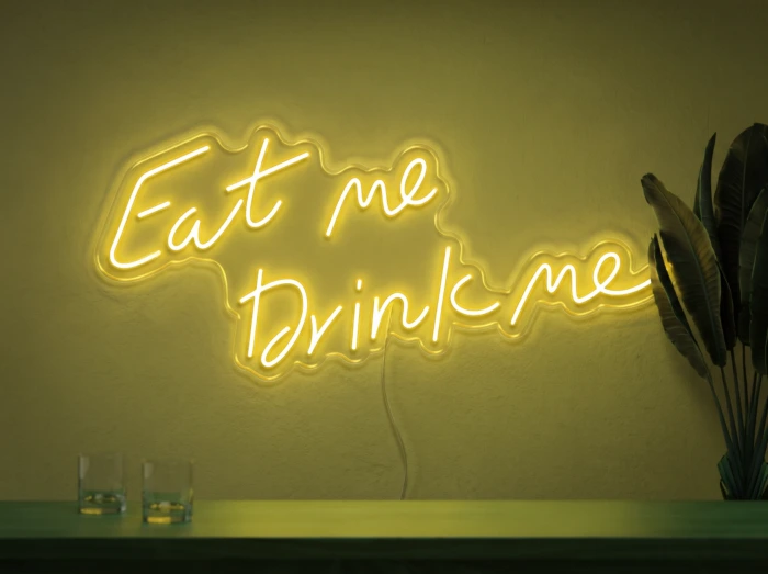 Neonskylt «Eat Me Drink Me» i gul LED-neon, monterad på vägg
