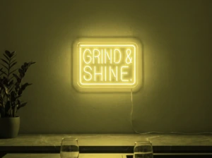 Neonskylt «Grind & Shine» i gul LED-neon, monterad på vägg