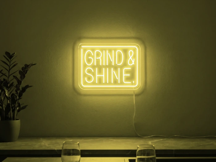 Neonskylt «Grind & Shine» i gul LED-neon, monterad på vägg