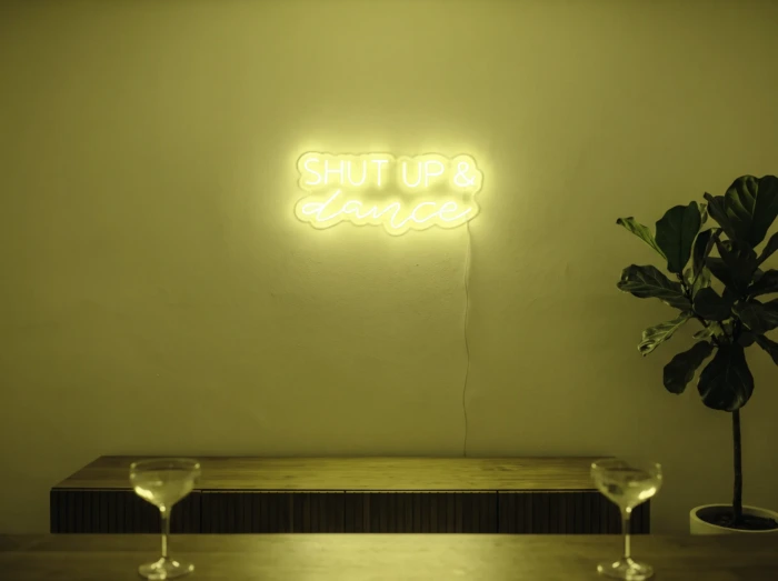 Neonskylt «Shut Up And Dance» i gul LED-neon, monterad på vägg