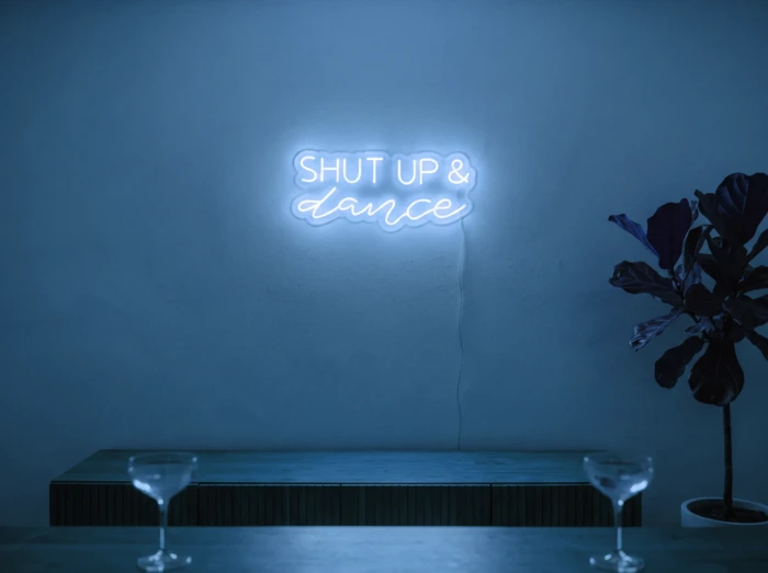Neonskylt «Shut Up And Dance» i isblå LED-neon, monterad på vägg